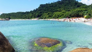 Una playa con aguas turquesas cristalinas, un afloramiento rocoso en primer plano y una colina cubierta de vegetación densa al fondo.