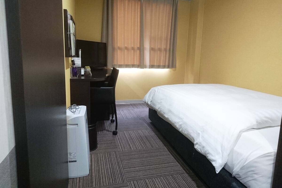A hotel room with a bed, a desk, a chair, and a small trash bin.
