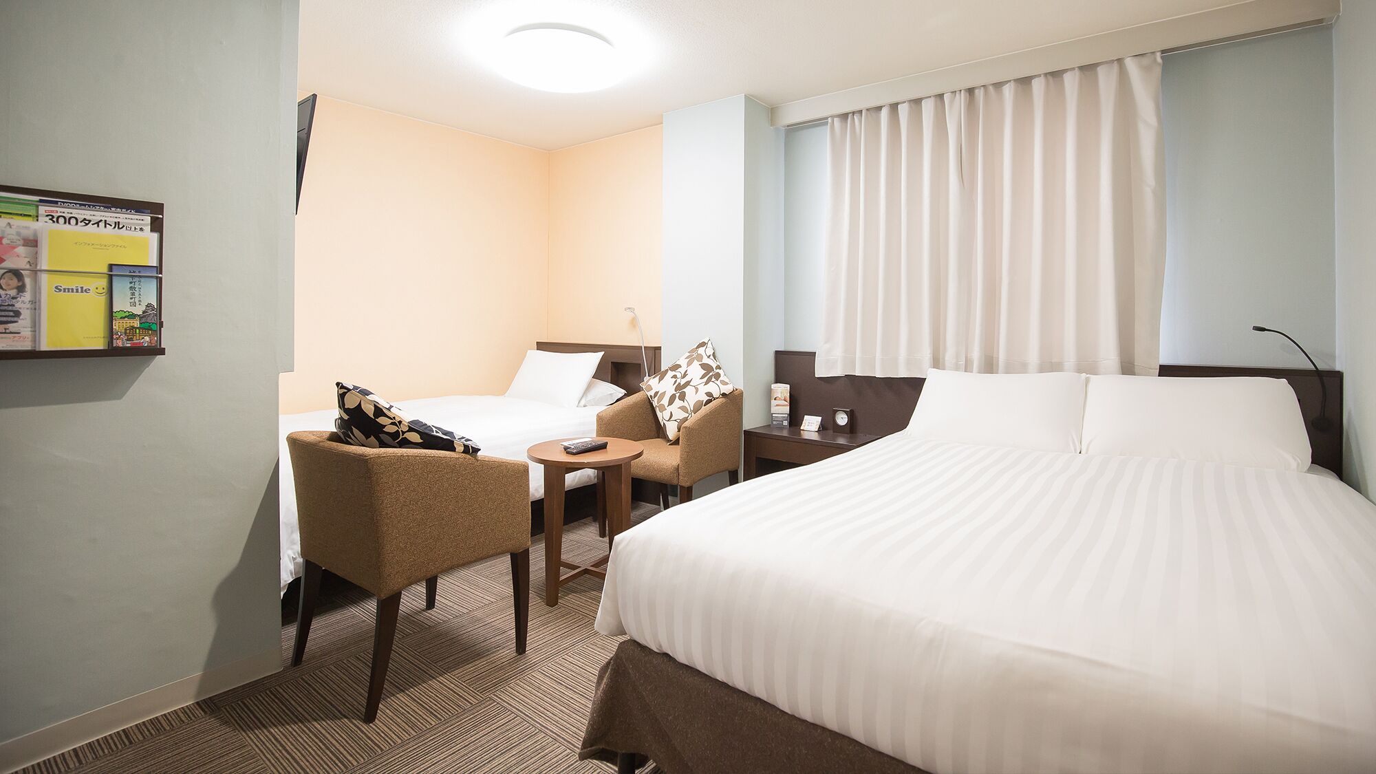 Habitación de hotel con cama, una silla, una mesita, una mesita de noche y una lámpara fijada a la pared.