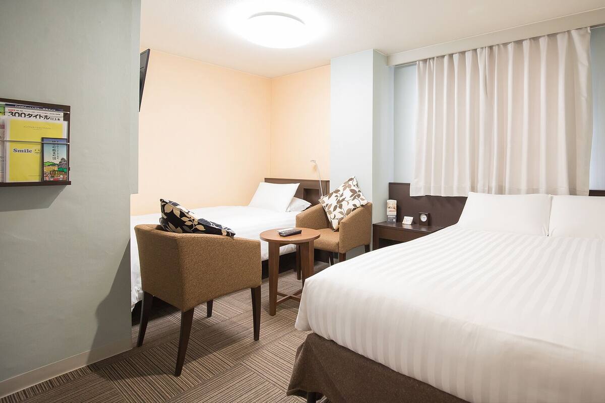 A hotel room with a bed, a chair, a small table, a nightstand, and a wall-mounted lamp.