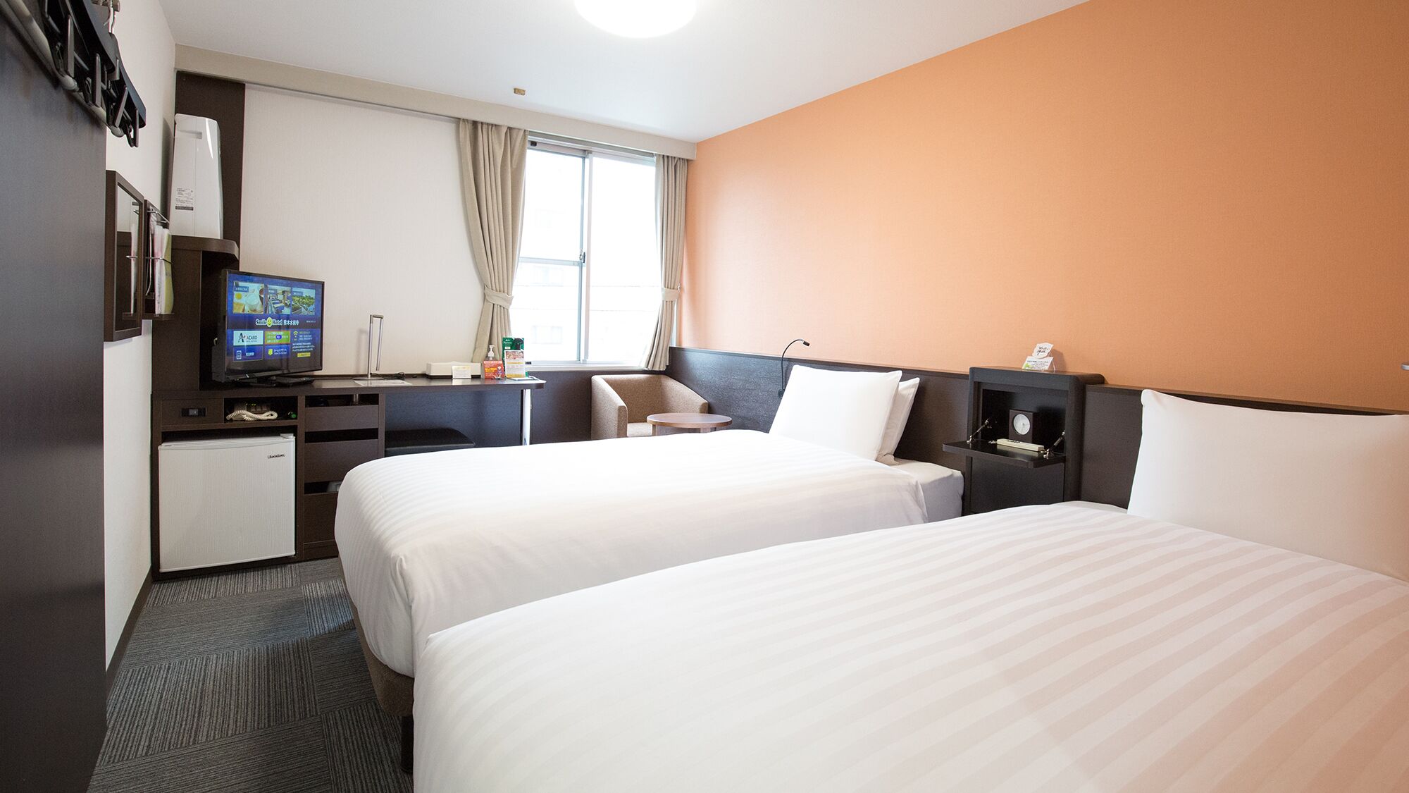 Een hotelkamer met twee bedden, een bureau met een computer, een kleine koelkast en een raam met gordijnen.