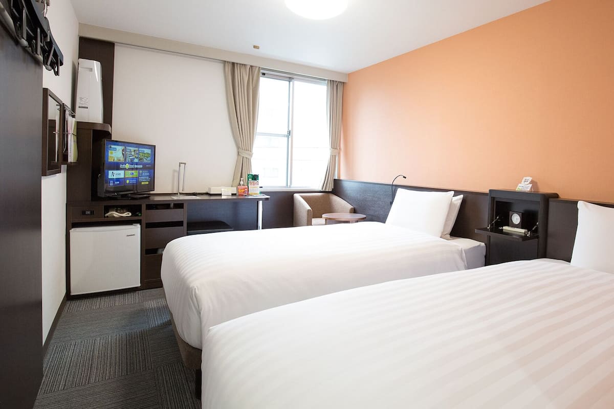 Habitación de hotel con dos camas, un escritorio con computadora, un refrigerador pequeño y una ventana con cortinas.