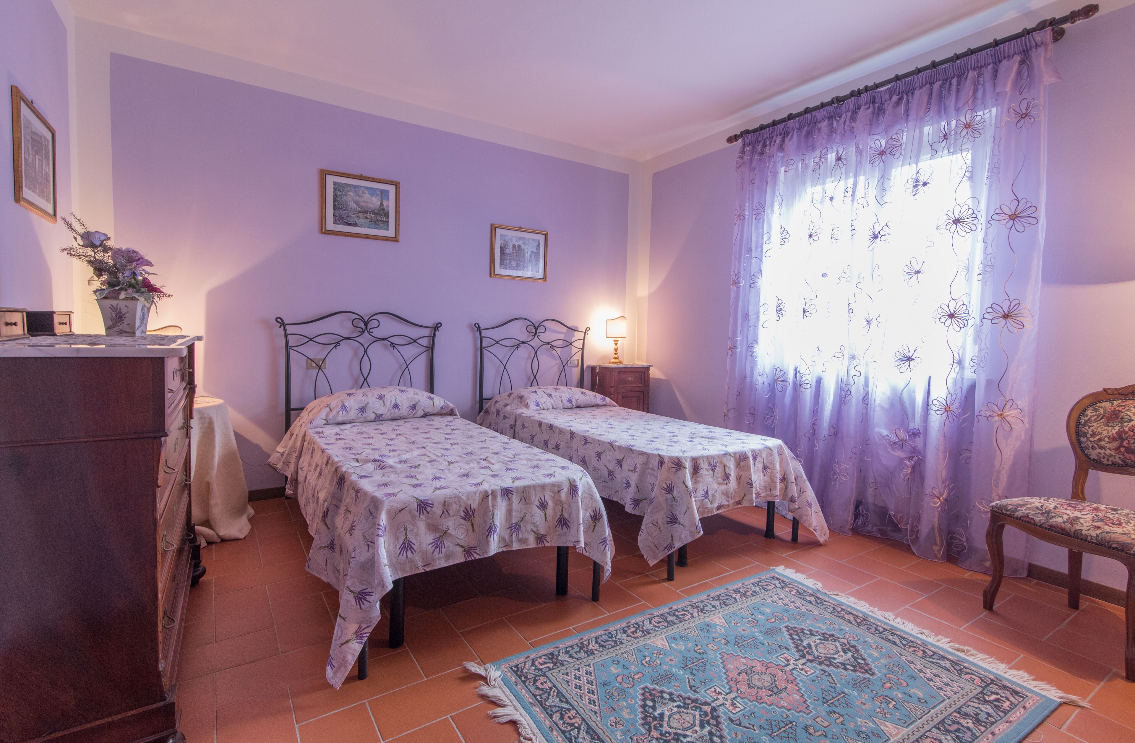 Une chambre avec deux lits simples, une commode en bois, une chaise et une fenêtre ornée de rideaux à motifs floraux violets.
