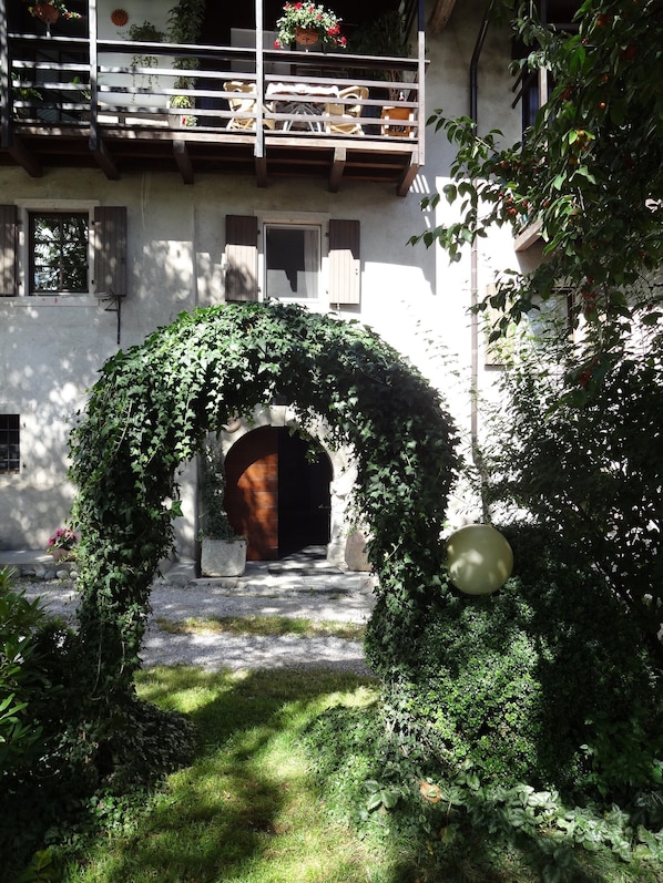A courtyard with a stone archway covered in ivy, a wooden balcony with potted plants, and a building with windows and shutters.