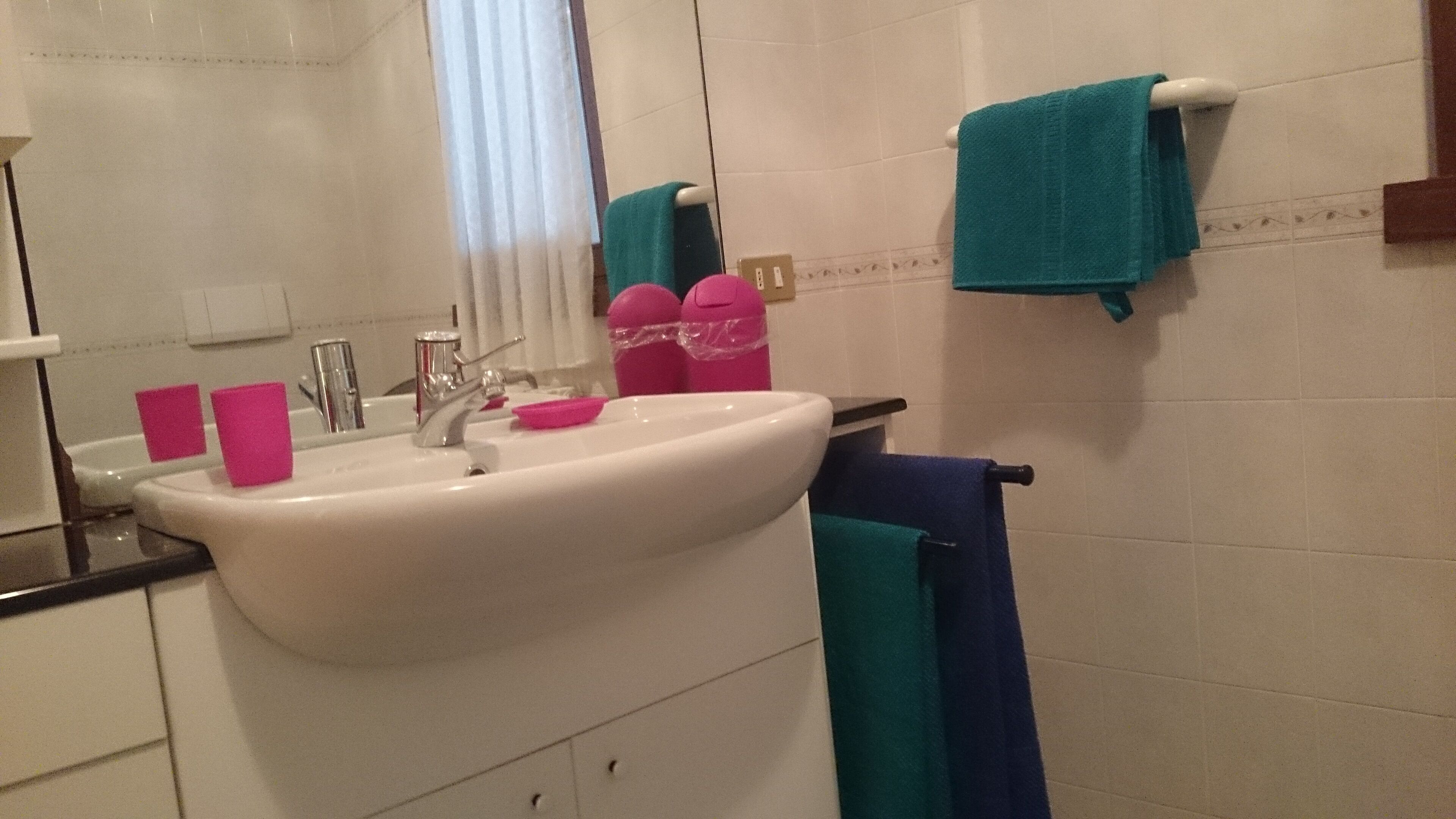Un baño con un lavamanos blanco, un espejo, una ventana con cortinas blancas y un perchero con una toalla azul.