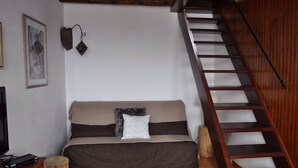 Un espacio de sala acogedor con un sofá, una escalera de madera y una lámpara fijada a la pared.