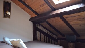 Un dormitorio con techo inclinado, vigas de madera, una cama con almohadas, un televisor sobre un mueble de madera y un cuadro enmarcado en la pared.