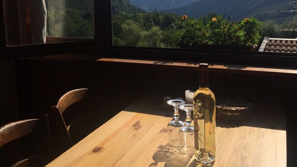 Una mesa de madera con una botella de vino, copas y una vela en una habitación con vistas a montañas a través de grandes ventanales.