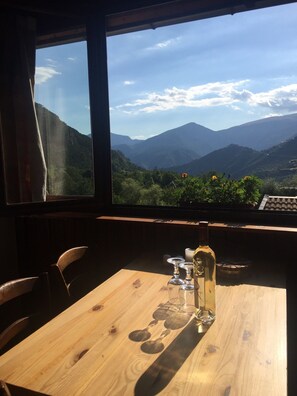 A wooden table with a bottle of wine, glasses, and a candle in a room with a view of mountains through large windows.