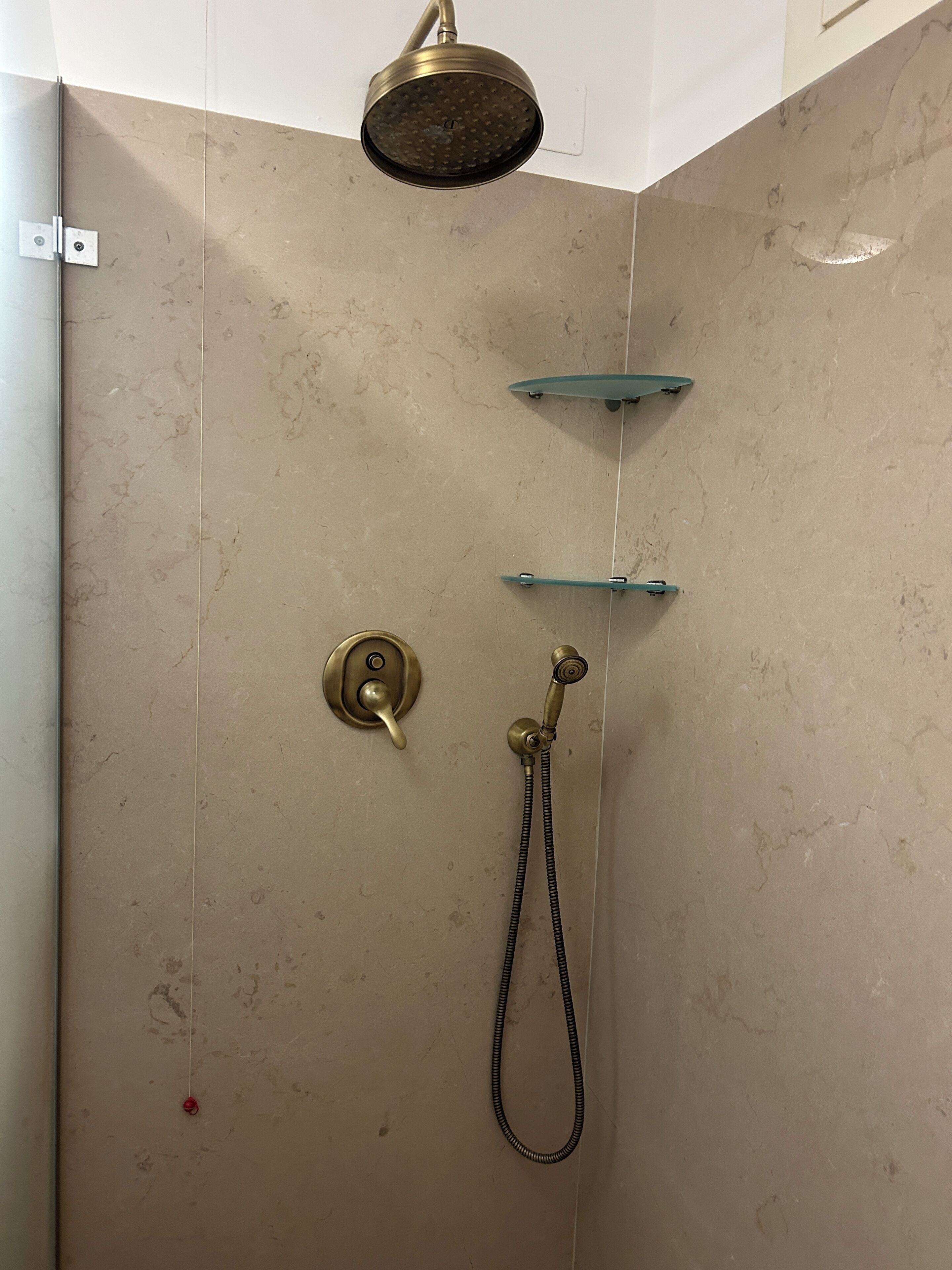 A shower with a handheld showerhead, a wall-mounted faucet, and a glass shelf.