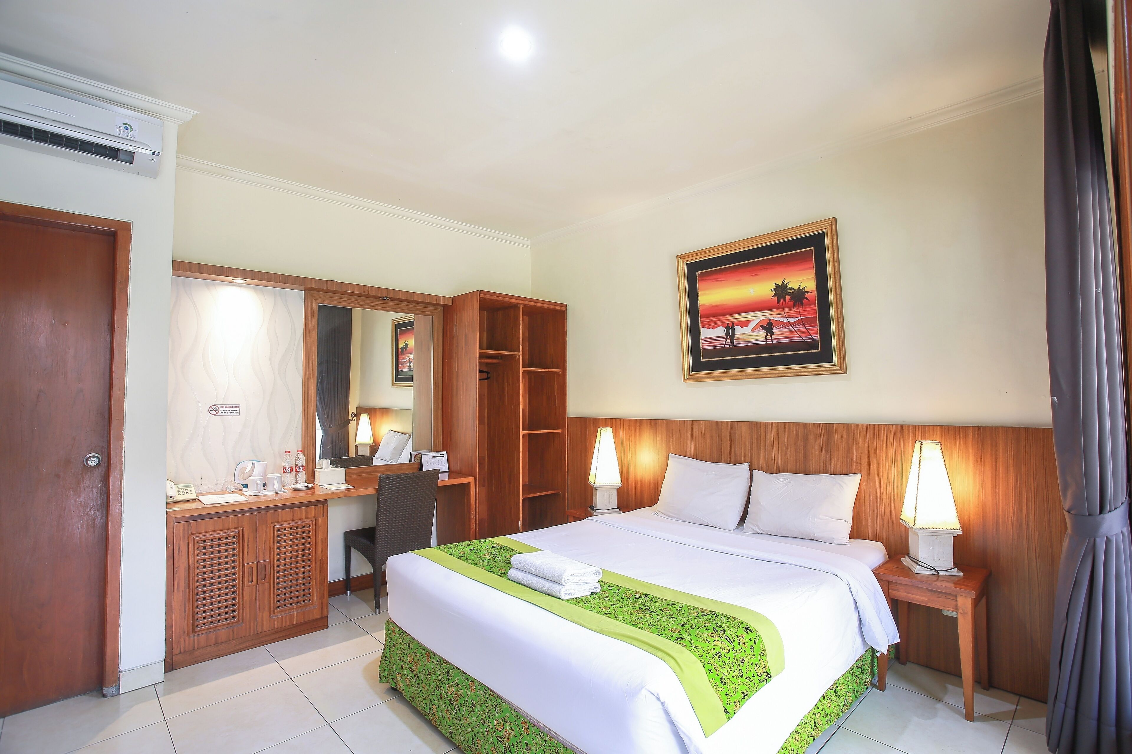 A hotel room with a bed, a desk, a wardrobe, a mirror, a painting, and a lamp.