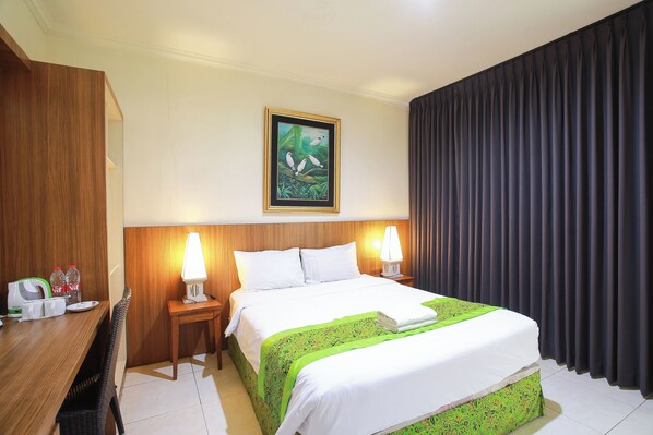 A hotel room with a bed, a desk, a chair, a lamp, and a painting on the wall. - Mirah Hotel (Banyuwangi)