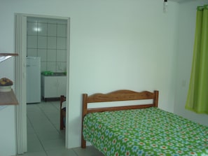Un dormitorio con una cama de madera, una colcha verde con motivos florales, una puerta blanca que da a una cocina y una cortina verde.