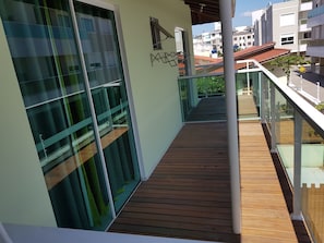 Un balcón moderno con barandillas de vidrio, piso de madera y vista a edificios cercanos.