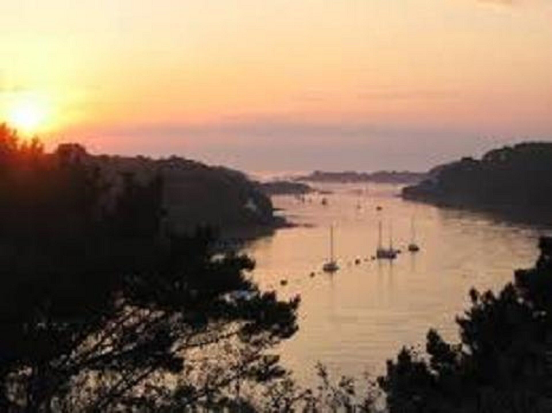 Une vue au coucher du soleil sur une rivière où l’on aperçoit des bateaux, le tout étant entouré d’arbres et de collines.