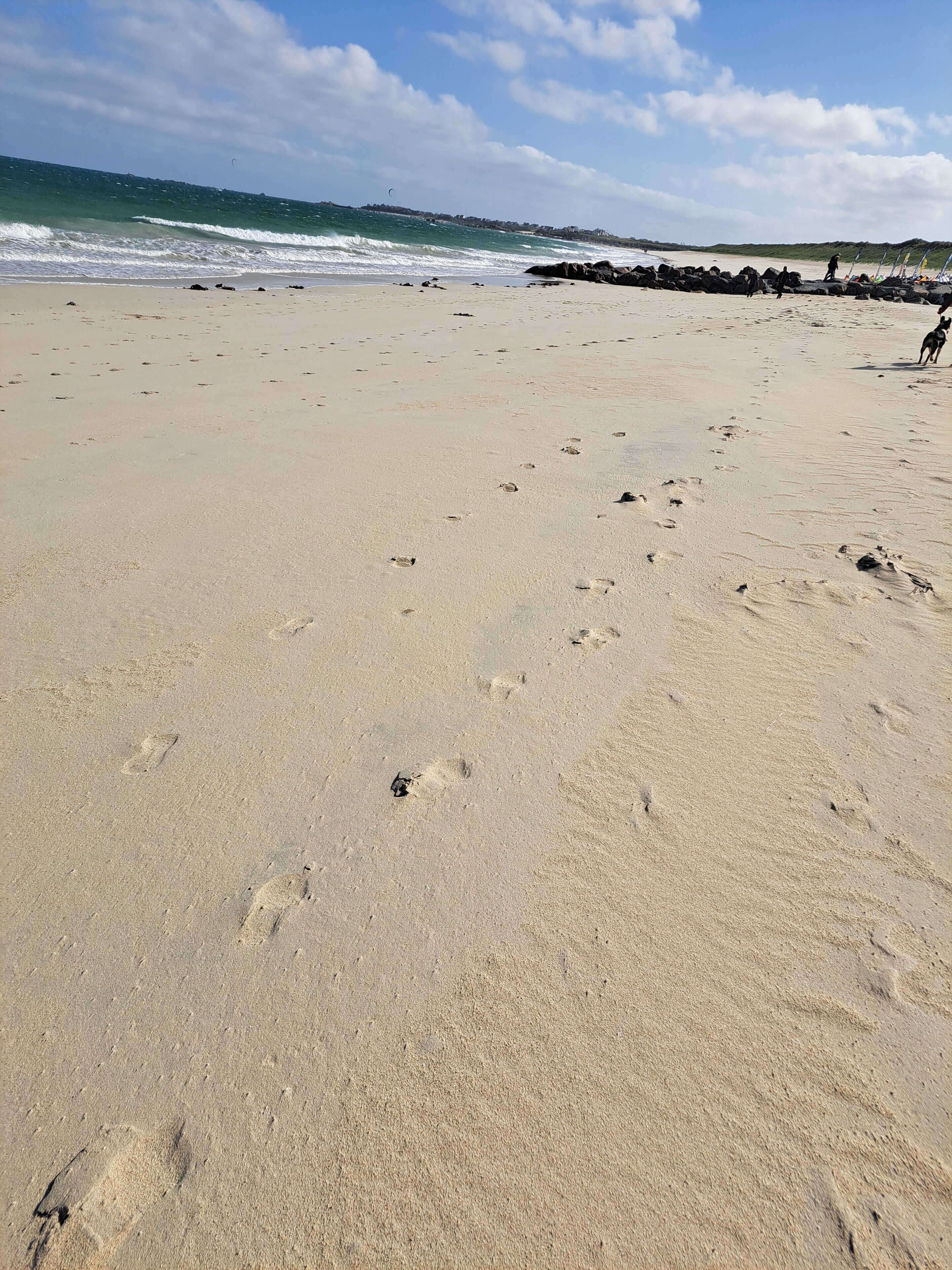 A sandy beach with footprints and a rocky breakwater in the background.