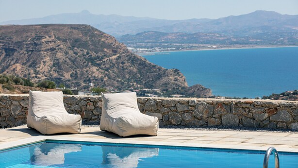 A poolside area with two beige lounge chairs, a stone wall, and a view of the sea and mountains.