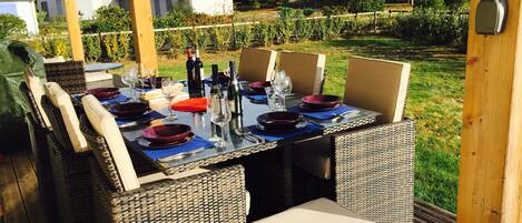 An outdoor dining area with wicker furniture, a glass table, and a view of the surrounding garden and houses.