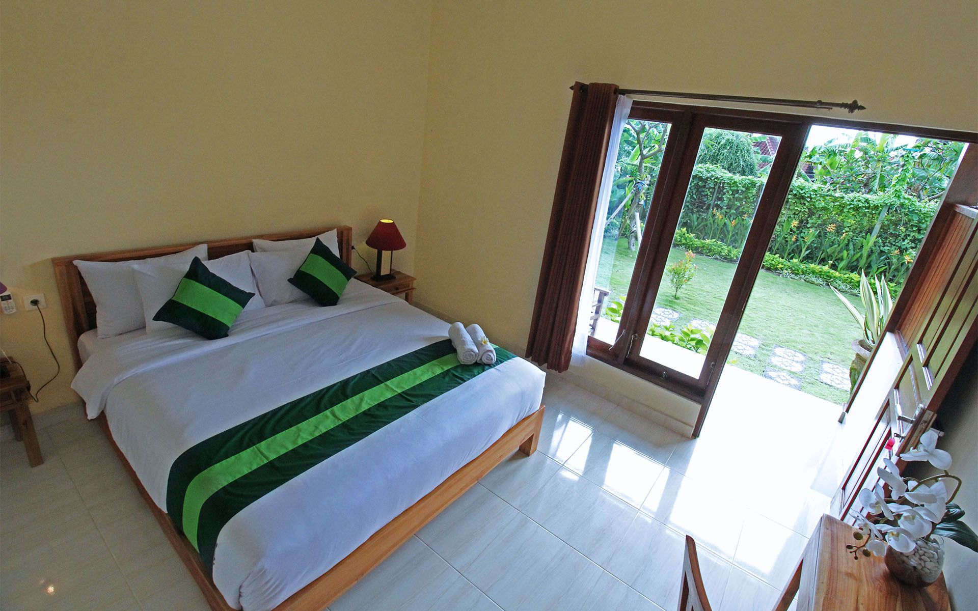 A hotel room with a bed, a nightstand, a lamp, and a view of a garden through a sliding door.