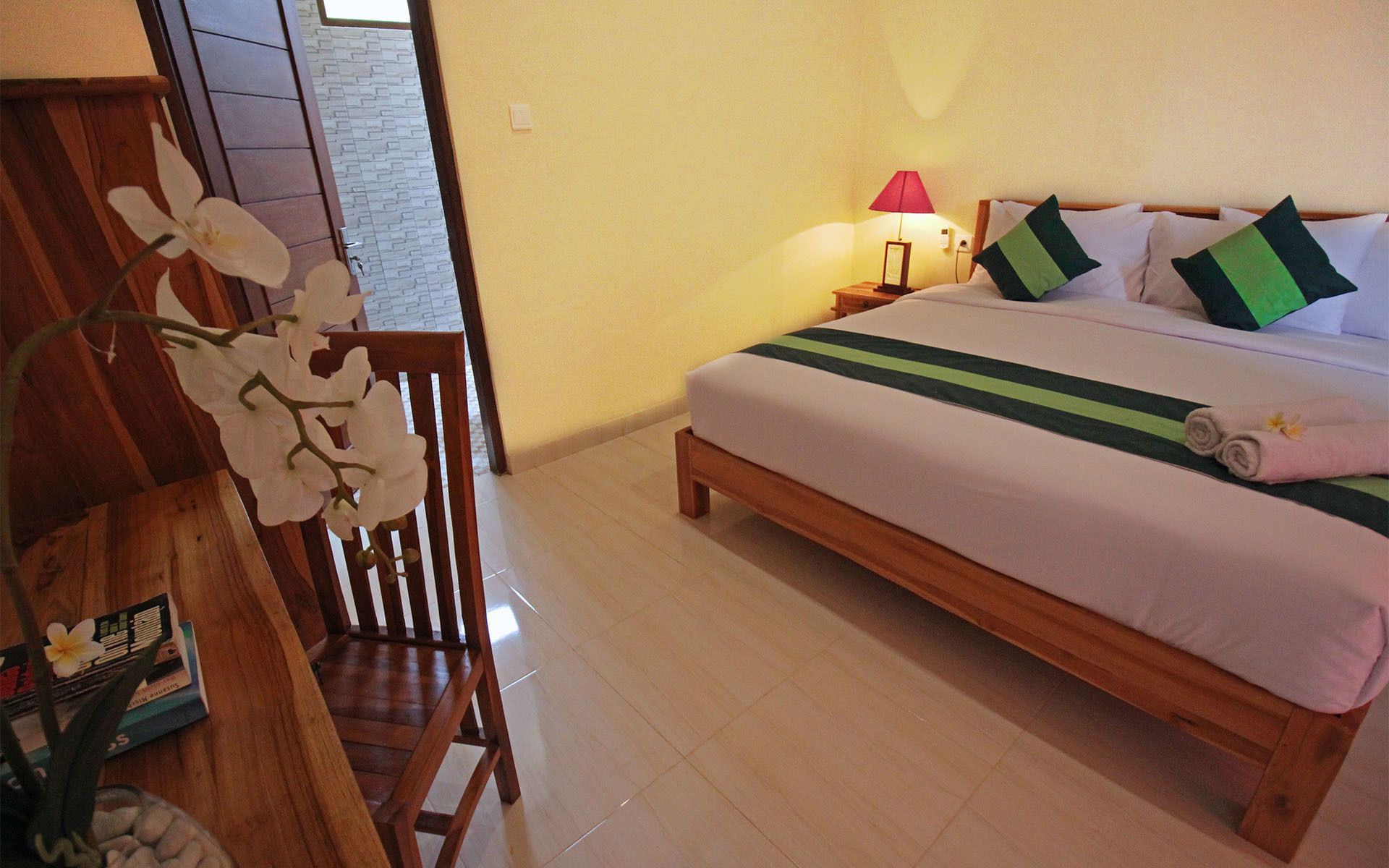 A hotel room with a wooden bed, a nightstand with a lamp, and a chair with an orchid plant.