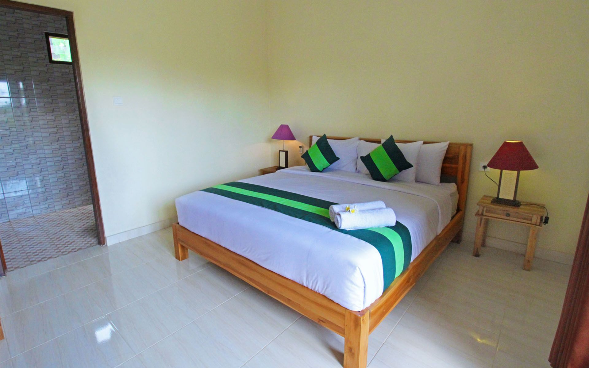 A neatly made bed with a wooden frame, a green and white striped bedspread, and a set of towels. There are two bedside lamps, one with a purple shade and one with a red shade. The room has a tiled floor and a brick-patterned wall.