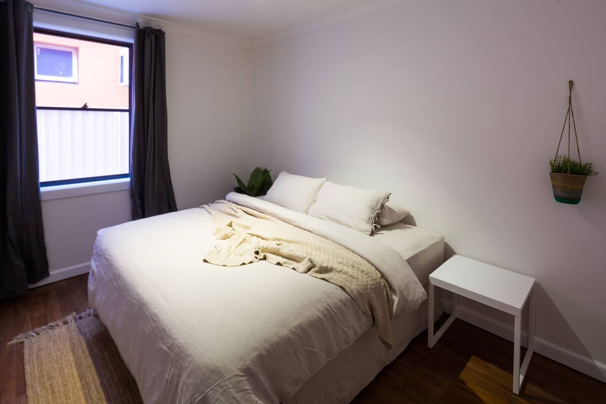 A neatly made bed with a white comforter and a beige throw blanket, a small bedside table, a hanging plant, and a window with curtains.