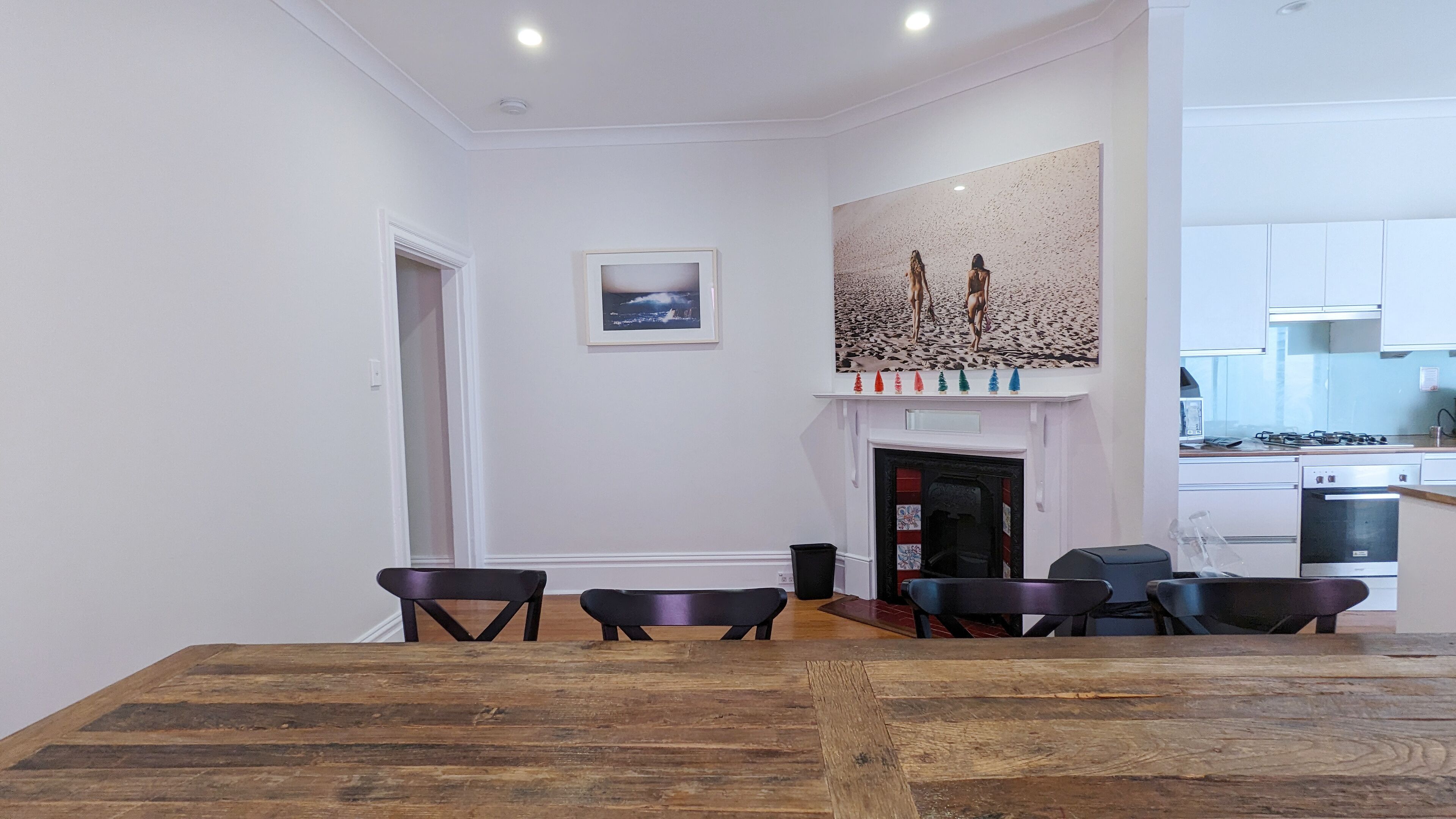 A dining area with a wooden table and chairs, a fireplace with a painting above it, and a kitchen in the background.