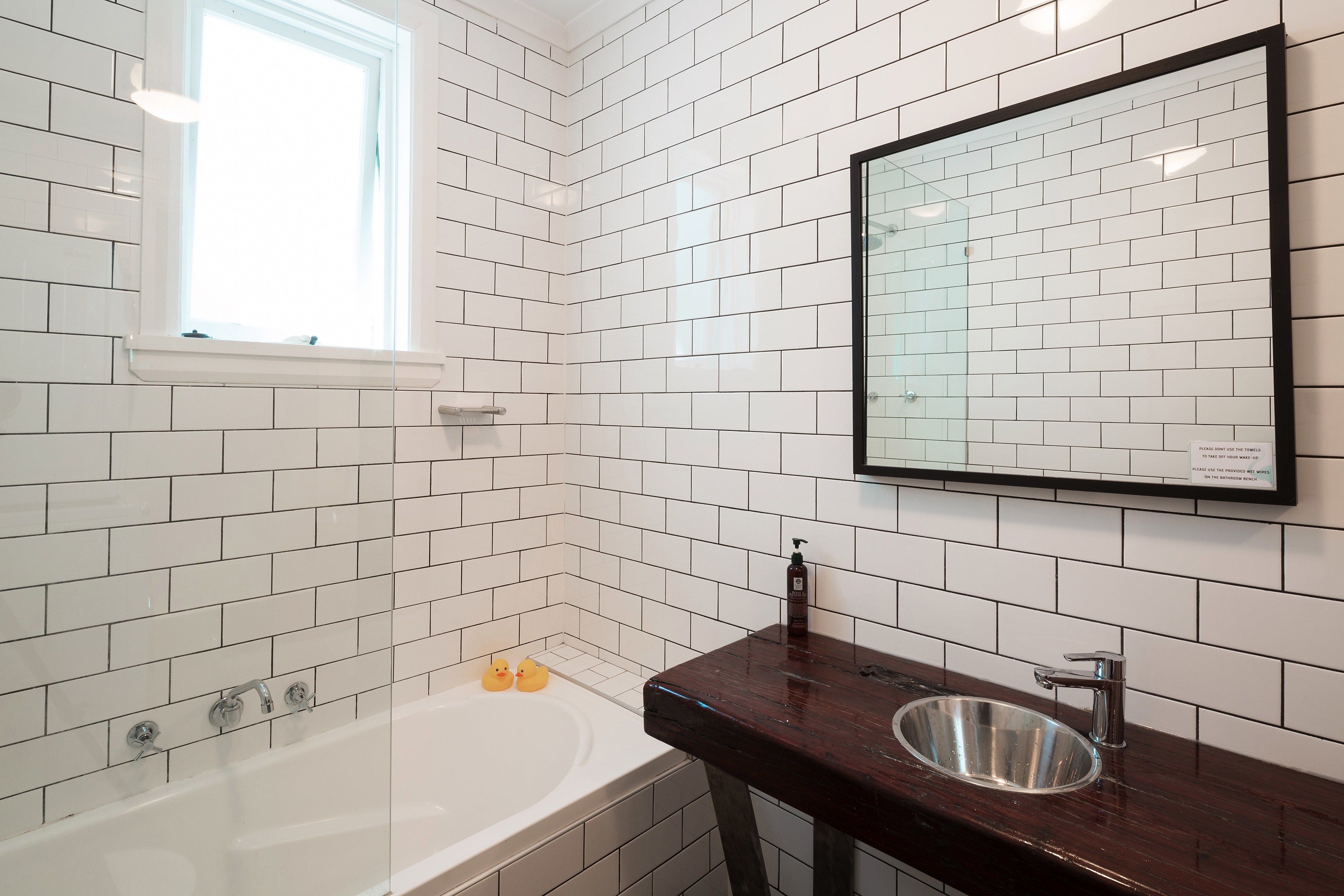 Un baño con paredes de azulejos blancos, una bañera, un lavabo con espejo y una ventana.