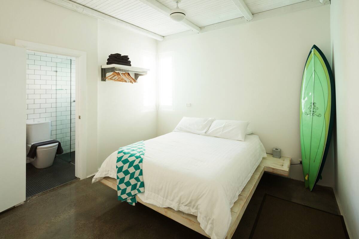 A bedroom with a bed, a green surfboard leaning against the wall, a small shelf with folded clothes, and a white door leading to a bathroom.