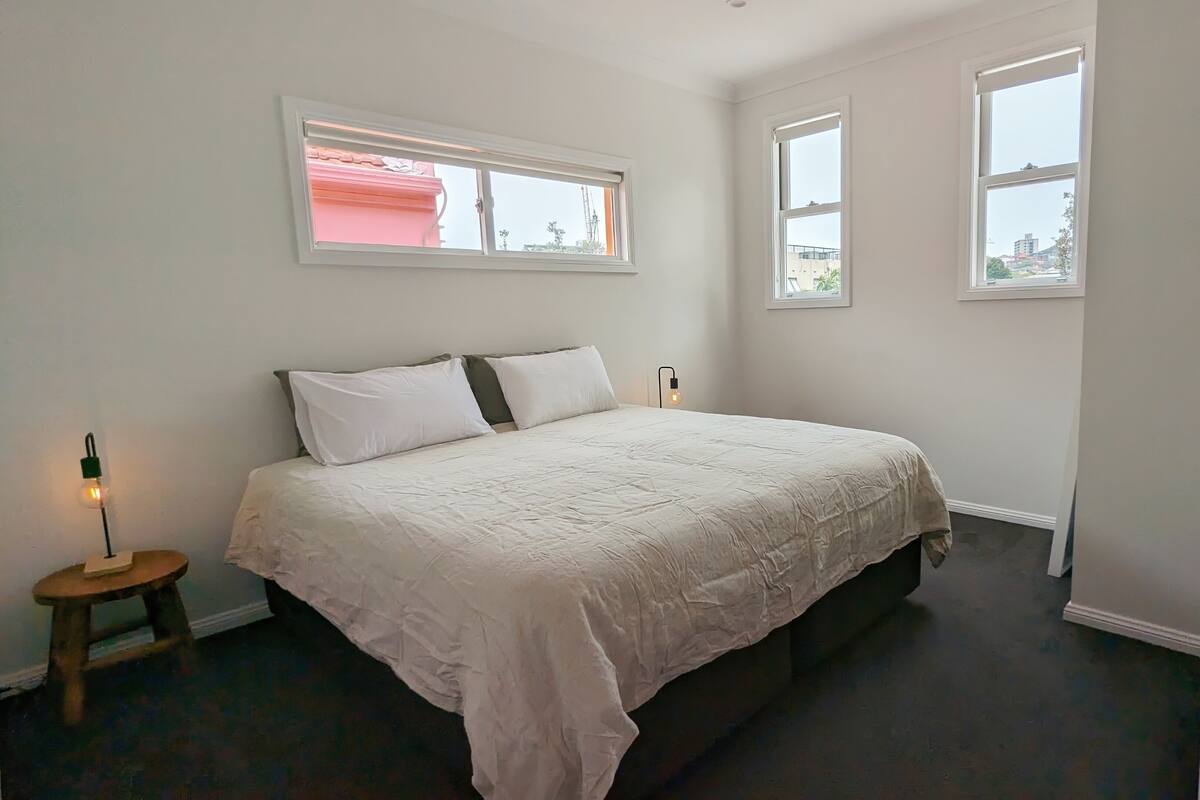 A bedroom with a bed, two bedside tables, and two windows.