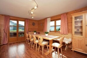 A wooden dining room with a table and chairs, large windows with curtains, and a cabinet.