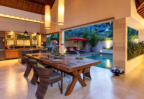 Un comedor con mesa de madera y sillas de mimbre, un área de descanso junto a la piscina con una sombrilla roja, y una cocina con vistas a la piscina.