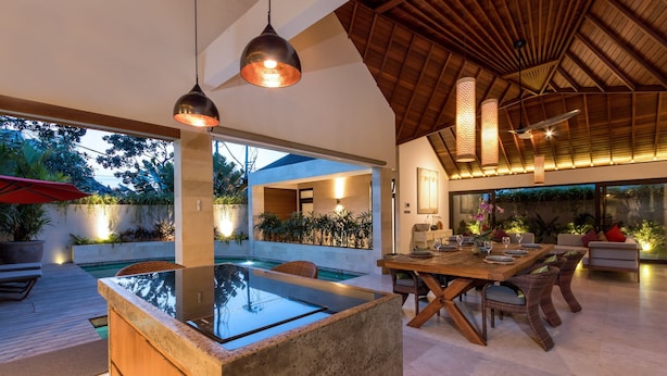 Un área de comedor exterior moderna con piscina, mobiliario de madera e iluminación colgante.