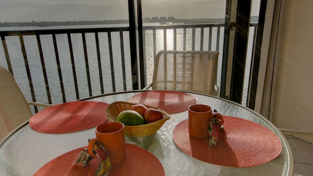A round glass table with red placemats, a fruit basket, and two orange mugs on a balcony overlooking a body of water. - Million Dollar View  Bay Sunrises & Gulf Sunsets-Feb and March open 2025 (Sarasota)