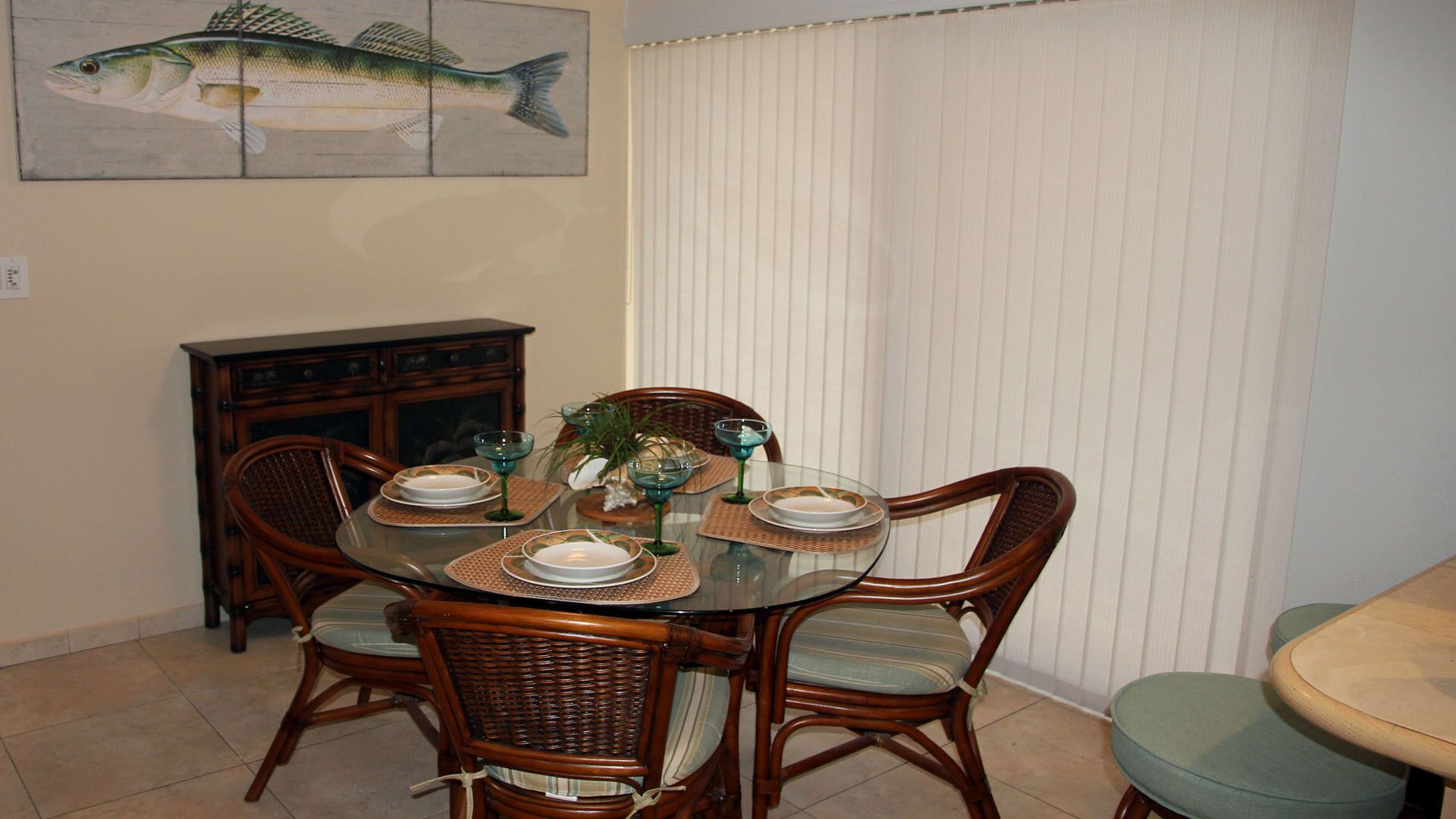 A dining area with a round glass table set for four, surrounded by wicker chairs with cushioned seats.