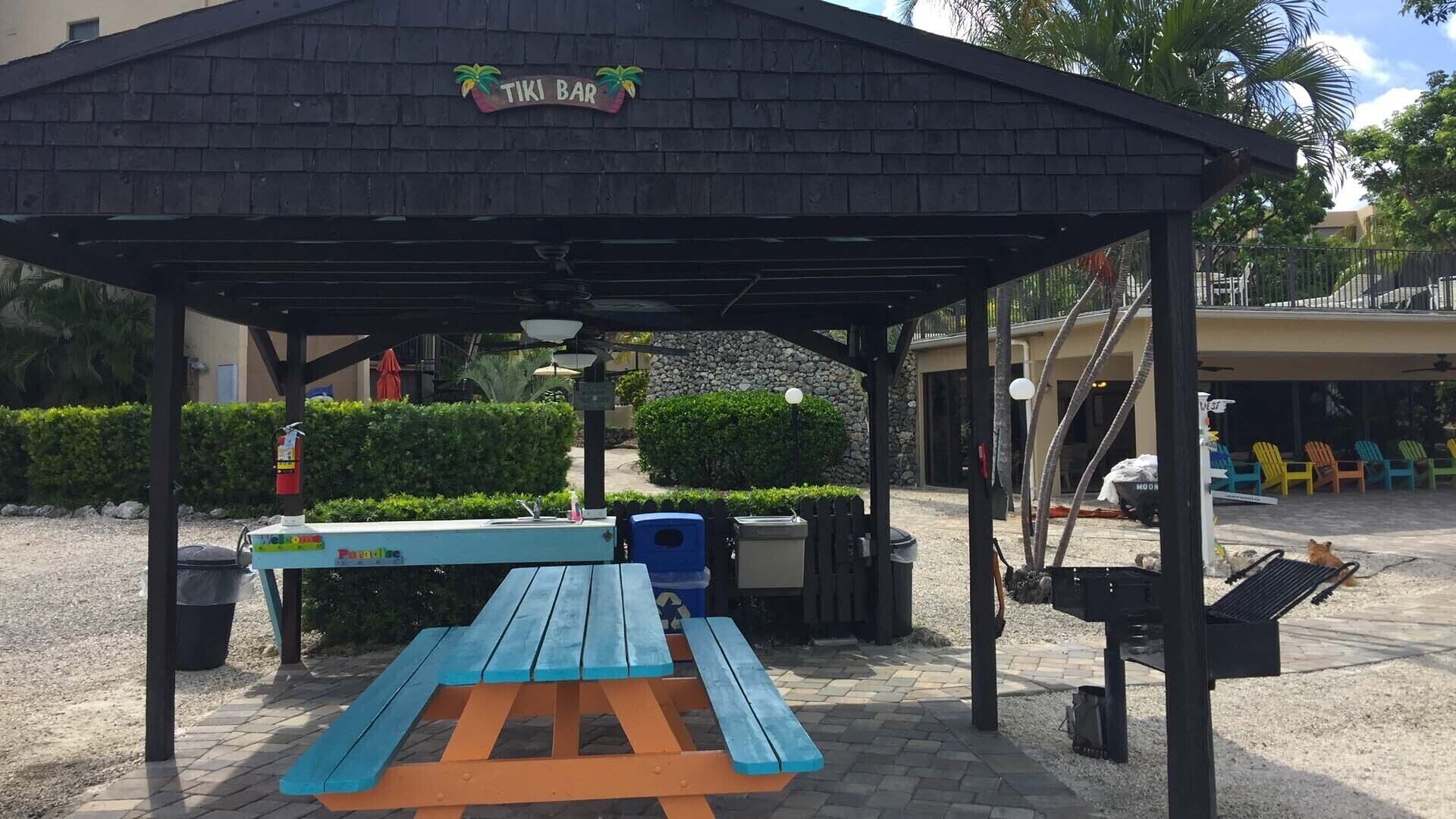 A covered picnic area with a bench and trash bins, a sign reading "TIKI BAR", and a building with colorful Adirondack chairs in the background.