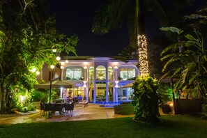 A modern house with a pool, surrounded by lush greenery and illuminated by string lights.