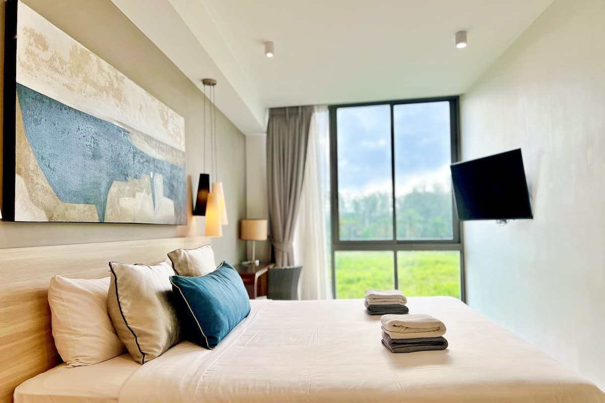 Une chambre d’hôtel avec un grand lit, un tableau au mur, un téléviseur fixé au mur et une fenêtre donnant sur un paysage verdoyant.