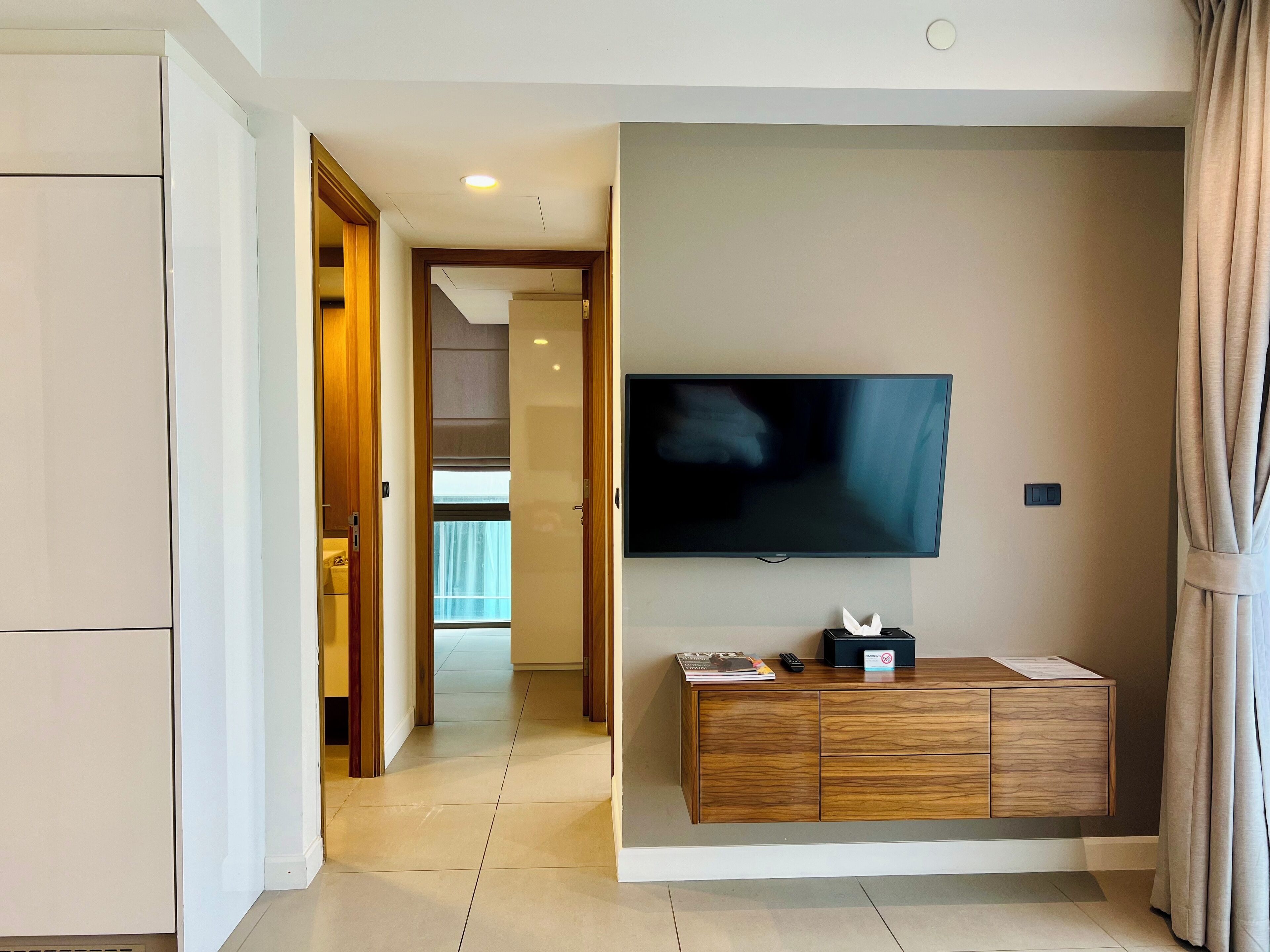 A hotel room with a flat-screen TV mounted on the wall, a wooden cabinet below it, and a door leading to another room.