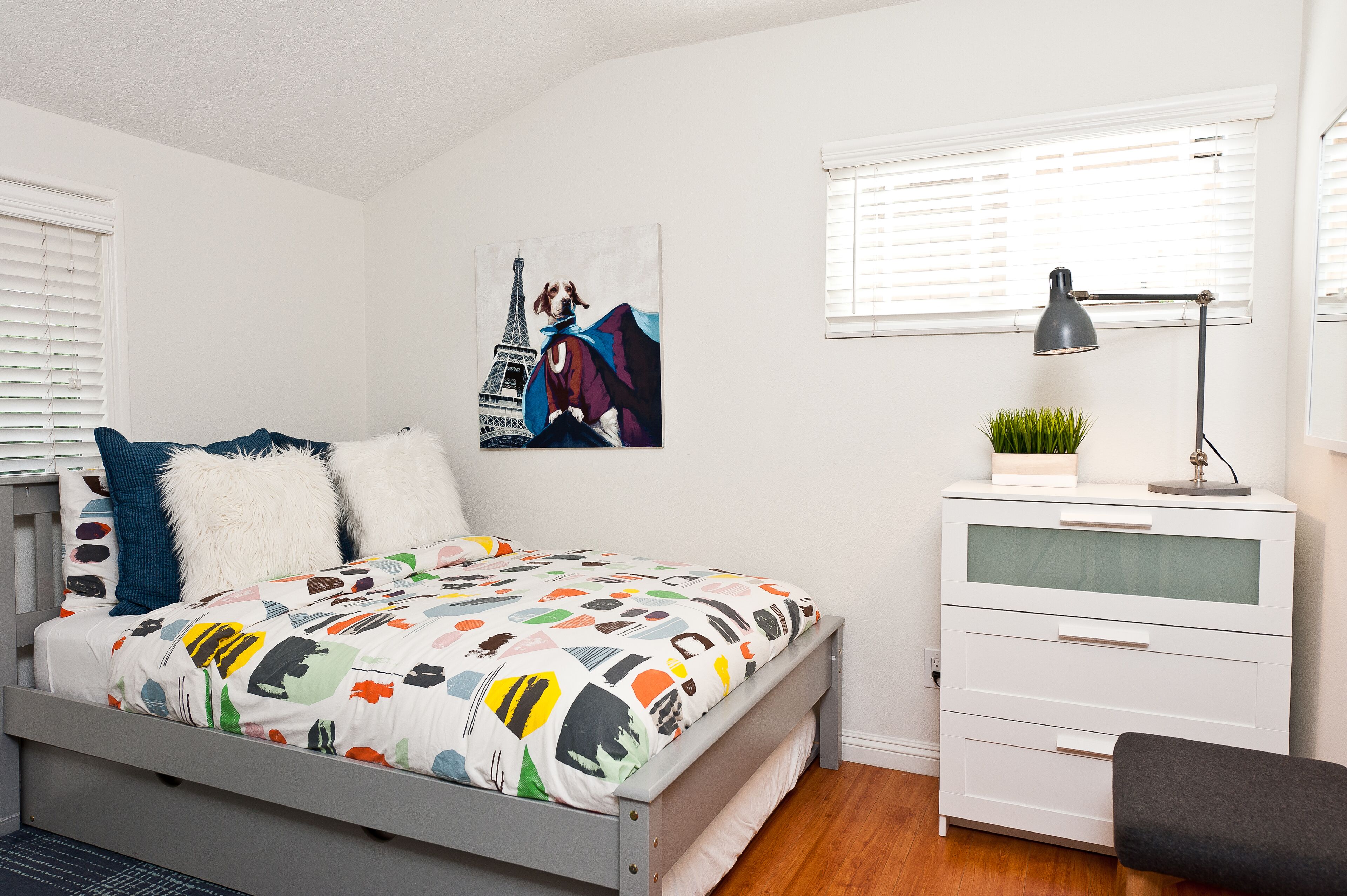 Une chambre à coucher avec un lit, une commode blanche, une lampe de bureau et un tableau représentant un chien portant une cape.