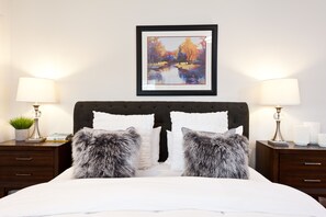 Un lit bien fait, avec une tête de lit sombre, deux oreillers moelleux et un tableau encadré au-dessus.