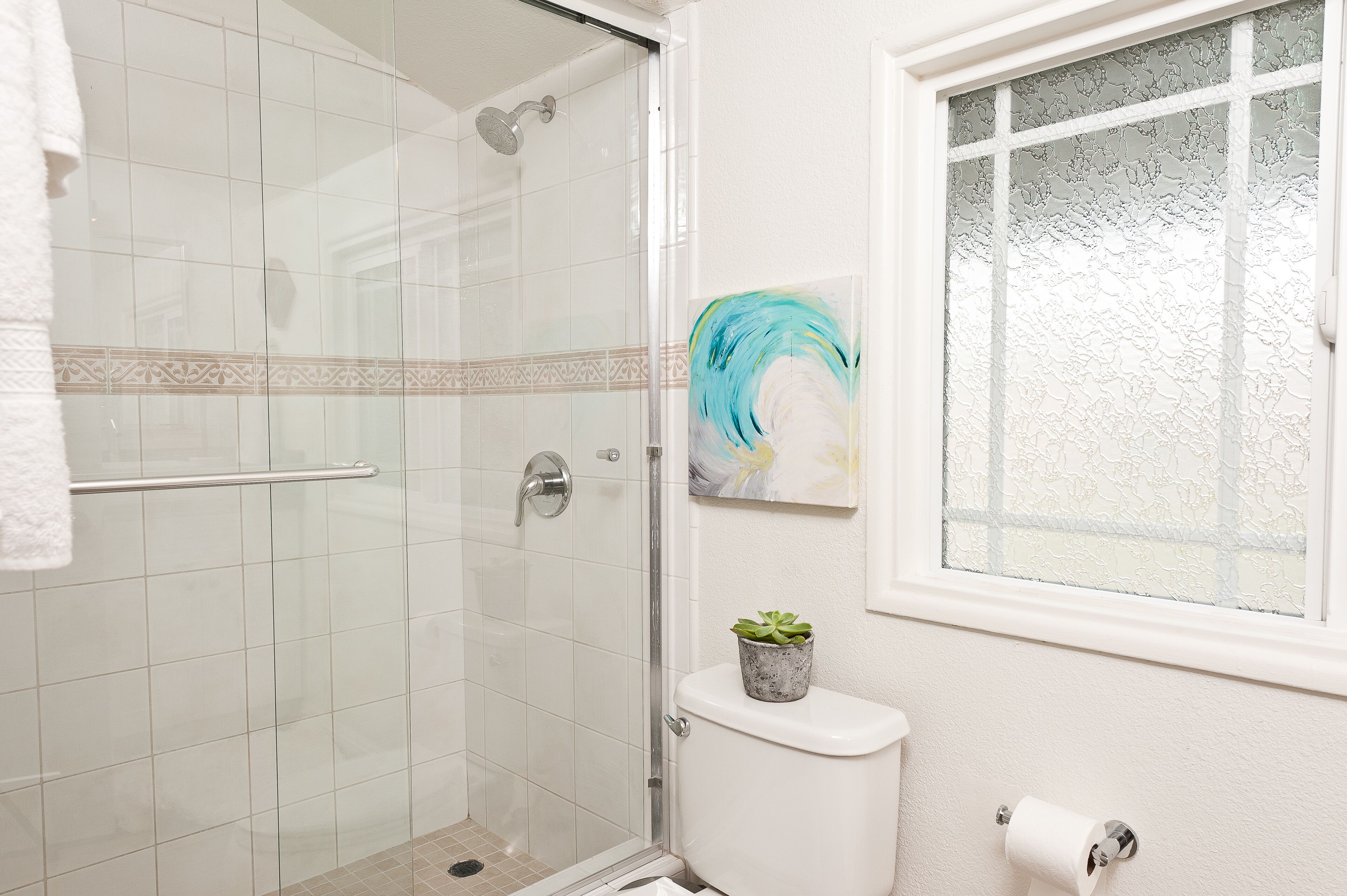 A bathroom with a glass shower enclosure, a toilet, a window with frosted glass, and a small potted plant on the toilet tank.