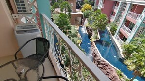 Un balcon doté d’une table et de chaises en verre, donnant sur une piscine agrémentée d’une cascade, le tout entouré d’une végétation luxuriante et surplombé par un immeuble à plusieurs étages également doté de balcons.