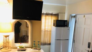 A compact hotel room with a kitchenette, a flat-screen TV mounted above the counter, a refrigerator, and a microwave. There is a chair and a bed with a colorful quilt. - PRIME STUDIO SAVE. UPSCALE BEACHFRONTS. SUN,SURF&SAND, TOWN 2 BLOCKS, PKG,CHAIRS (Madeira Beach)