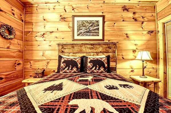 A cozy cabin-style room with a wooden interior, a bed with a patterned quilt, a framed picture on the wall, and a small table with a lamp.