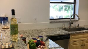 A kitchen with granite countertops, a bottle of California Roots Moscato, a cutting board with vegetables, and a window with a view of trees. - A little piece of heaven on the river (Port St Lucie)