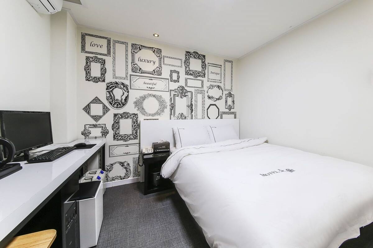 Habitació d'hotel amb un llit, un escriptori amb un ordinador i una paret decorada amb quadres negres i blancs emmarcats.
