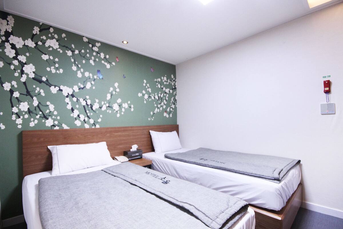 Habitació d'hotel amb dos llits individuals, un amb fusta i un panell mural floral a la paret verd.