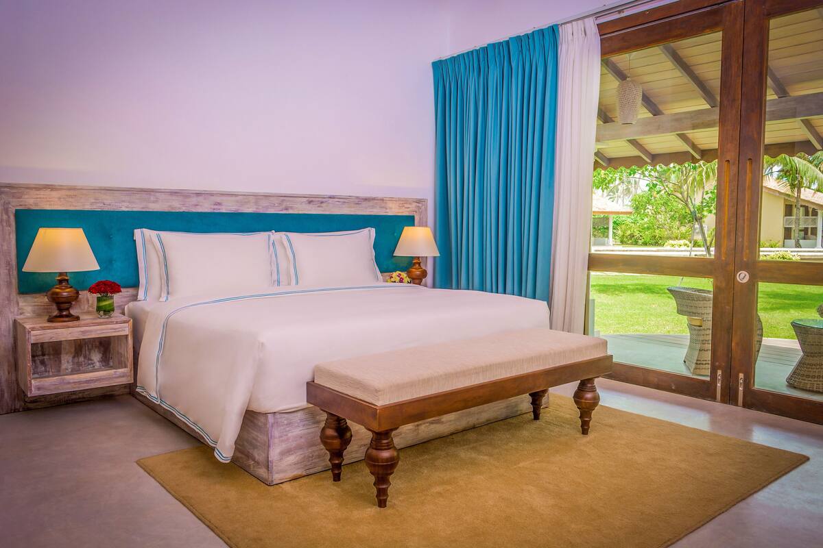 Una cama con cabecera de madera, dos mesitas de noche con lámparas, un banco y vista a un jardín a través de un ventanal.