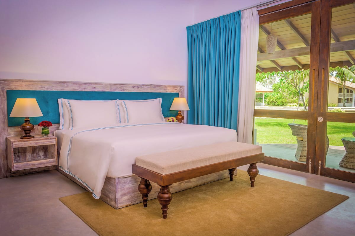 Una cama con cabecera de madera, dos mesitas de noche con lámparas, un banco y vista a un jardín a través de un ventanal.